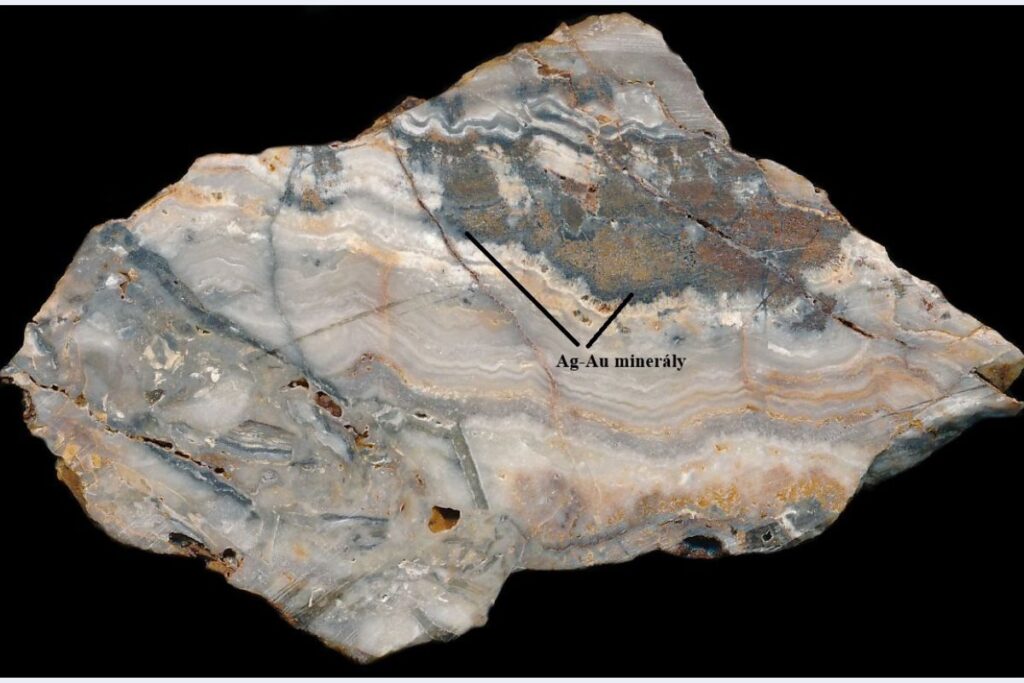 Typická páskovaná žilovina z lokality Šibeničný vrch so zhlukmi minerálov striebra (čierne) a zlatom. Šírka záberu je 9 cm.